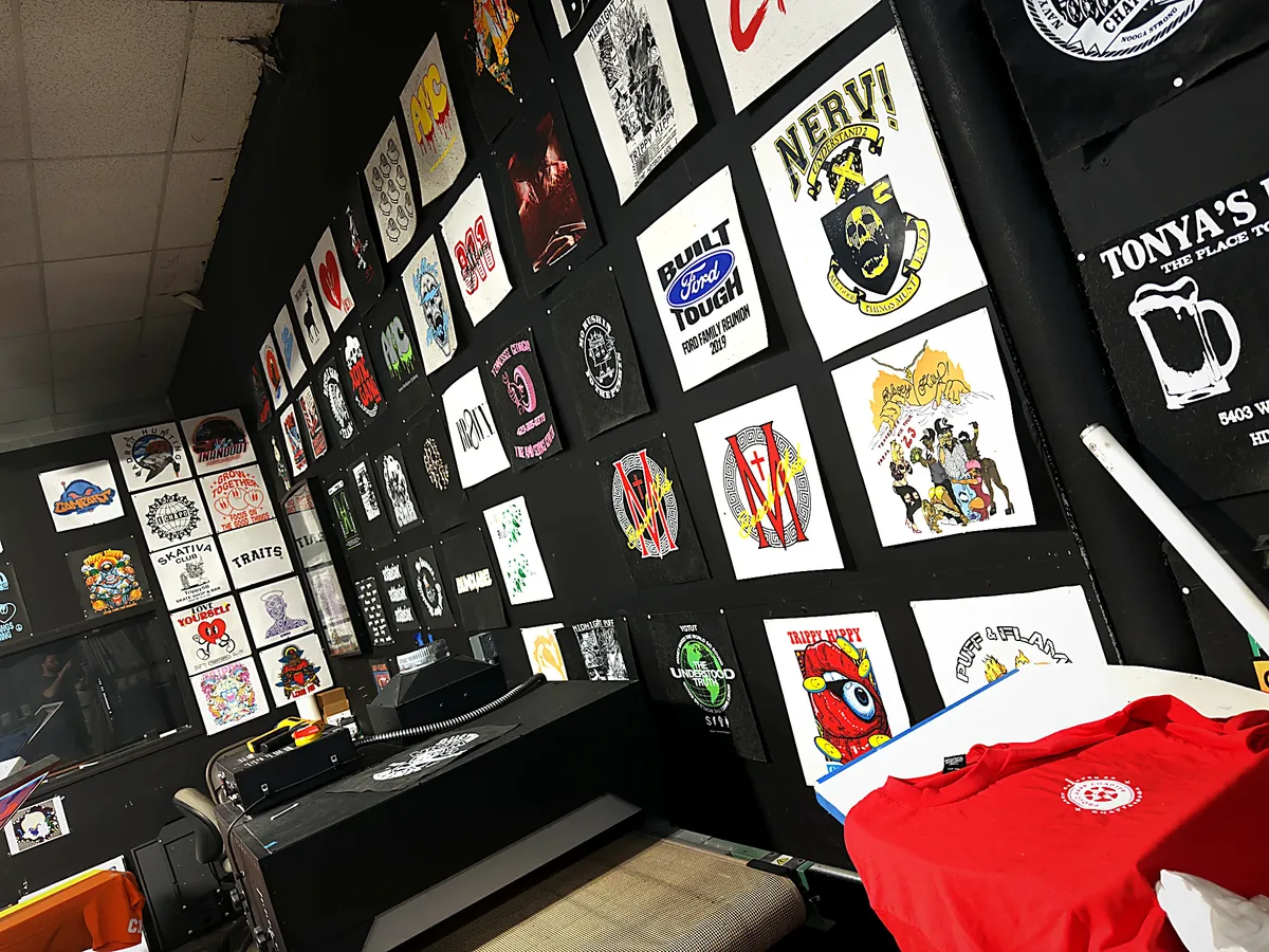 Wall of framed prints above the press, a red tee up next